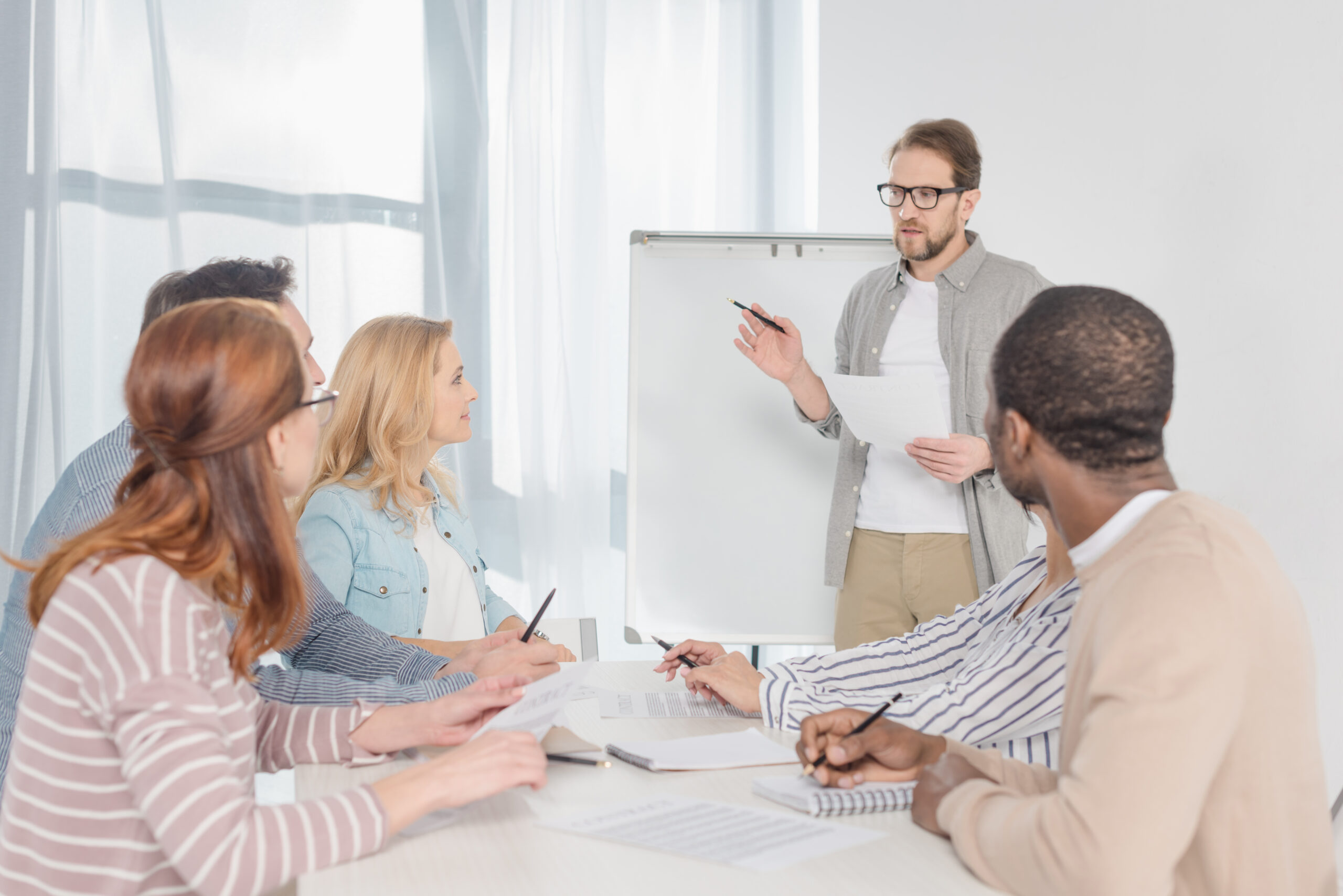 middle aged man in in eyeglasses with papers standing at whiteboard and having conversation with his